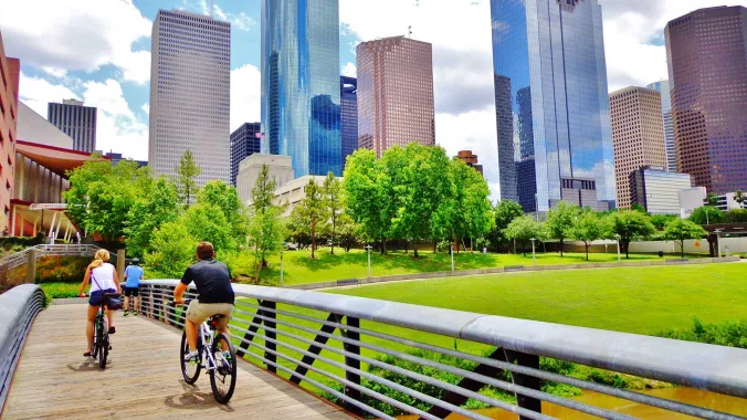 bicyclists cross bridge in park with houston skyline in background