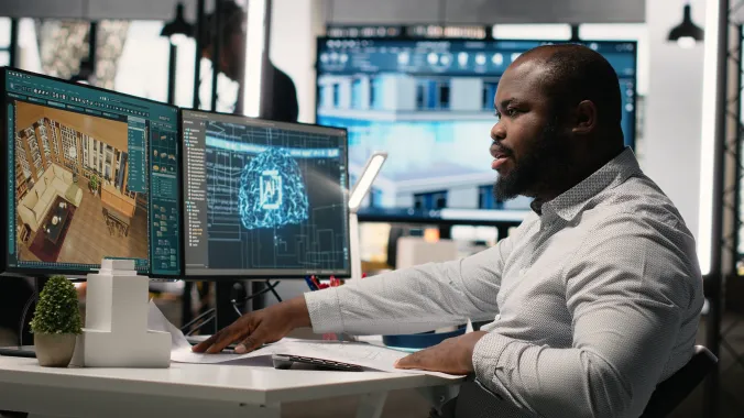 man sitting in front of two computer monitors with the keft side showing a floor plan and the right side showing an image of a blue light up brain with AI on it