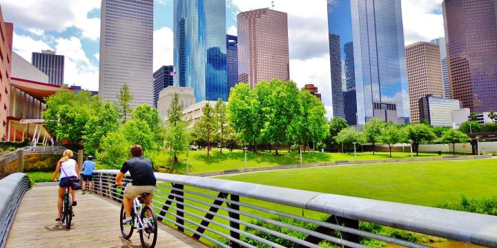 bicyclists cross bridge in park with houston skyline in background