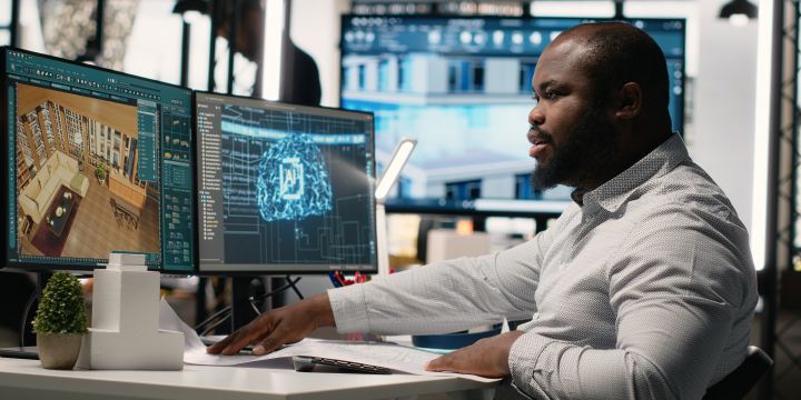 man sitting in front of two computer monitors with the keft side showing a floor plan and the right side showing an image of a blue light up brain with AI on it